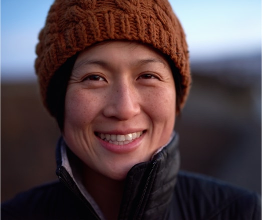 A person smiles warmly, wearing a brown knitted hat and a dark jacket, against a blurred outdoor background. The lighting is soft, adding to the serene and pleasant atmosphere of the scene.