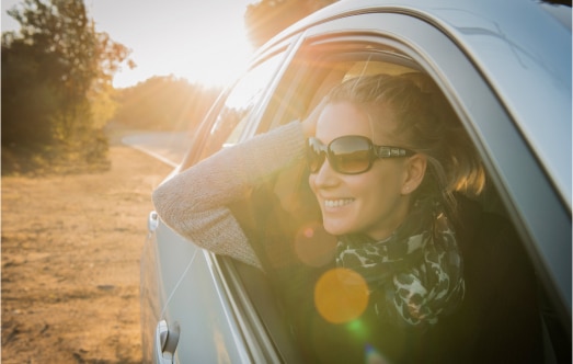 A person wearing sunglasses and a scarf leans out of a car window, smiling broadly. The sun is setting in the background, casting a warm glow and creating lens flares. The road and trees are visible beside the car.