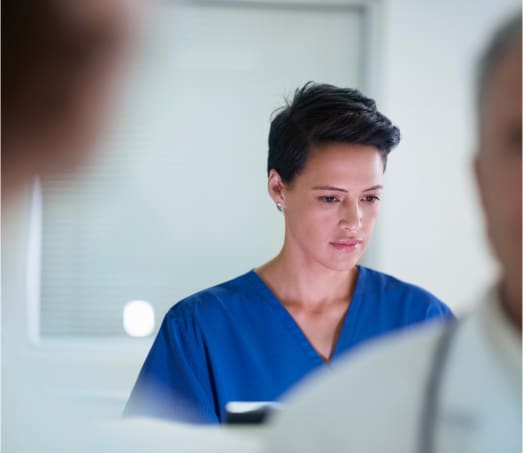 A medical professional with short dark hair, wearing blue scrubs, is focused on something in a bright room. They are in the background, while two blurred figures are seen closer to the camera, one wearing a stethoscope.