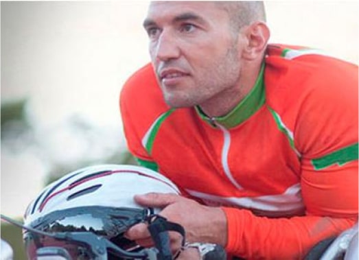 A cyclist in a bright red and green jersey, holding a white helmet, gazes intently forward. The background is blurred, emphasizing the focus on the cyclist.