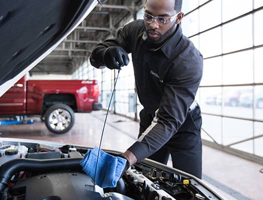 A mechanic, wearing safety goggles and gloves, checks the oil level of a car engine with a dipstick. He holds a blue cloth and stands in a garage with a red truck visible in the background.