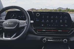 Interior view of a car dashboard featuring a Chevrolet steering wheel and a large central touchscreen displaying various apps. The background shows a scenic landscape with mountains and greenery under a cloudy sky.