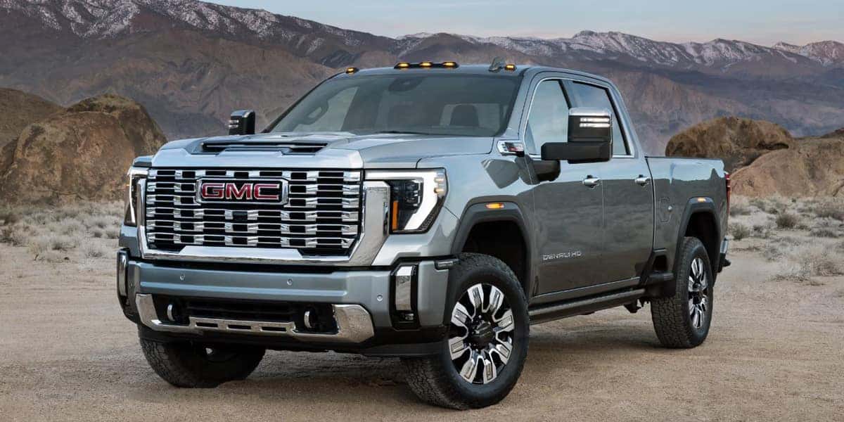 A silver GMC pickup truck is parked on a rocky terrain with mountains in the background. The truck features a large grille, prominent headlights, and a rugged design. The setting is under a clear sky, highlighting the vehicles robust build.