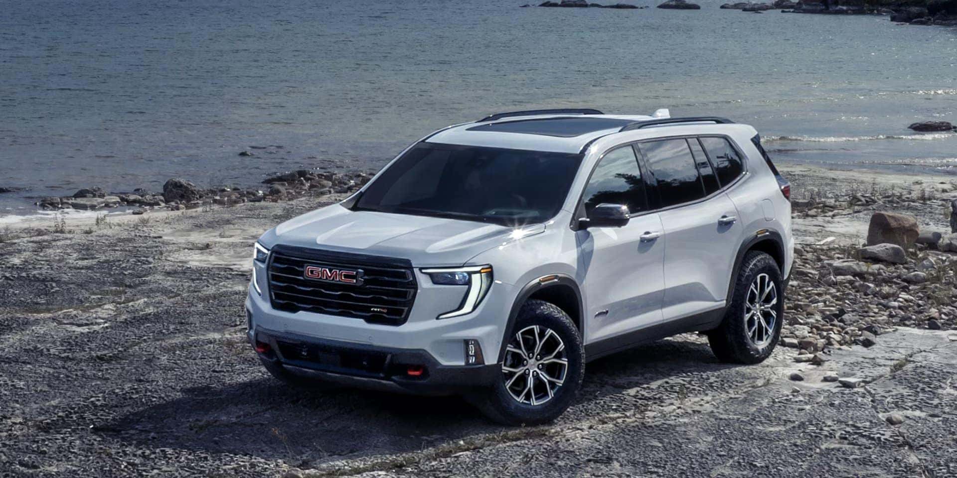 A white GMC SUV is parked on a rocky shoreline beside a calm body of water. The vehicle is under a clear sky, showcasing its sleek design and modern features, including prominent headlights and a bold grille. Rocks and water are visible in the background.