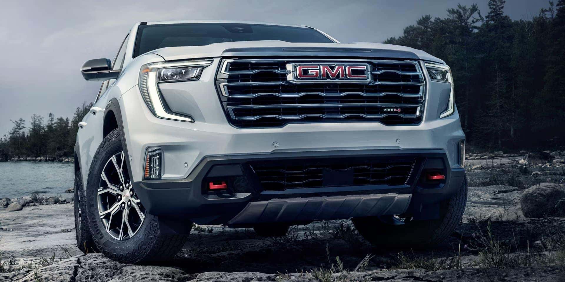 A white GMC truck with prominent grille and logo is parked on rocky terrain. The vehicle has black wheels and appears rugged, suitable for off-road. In the background, there are trees and a body of water under an overcast sky.
