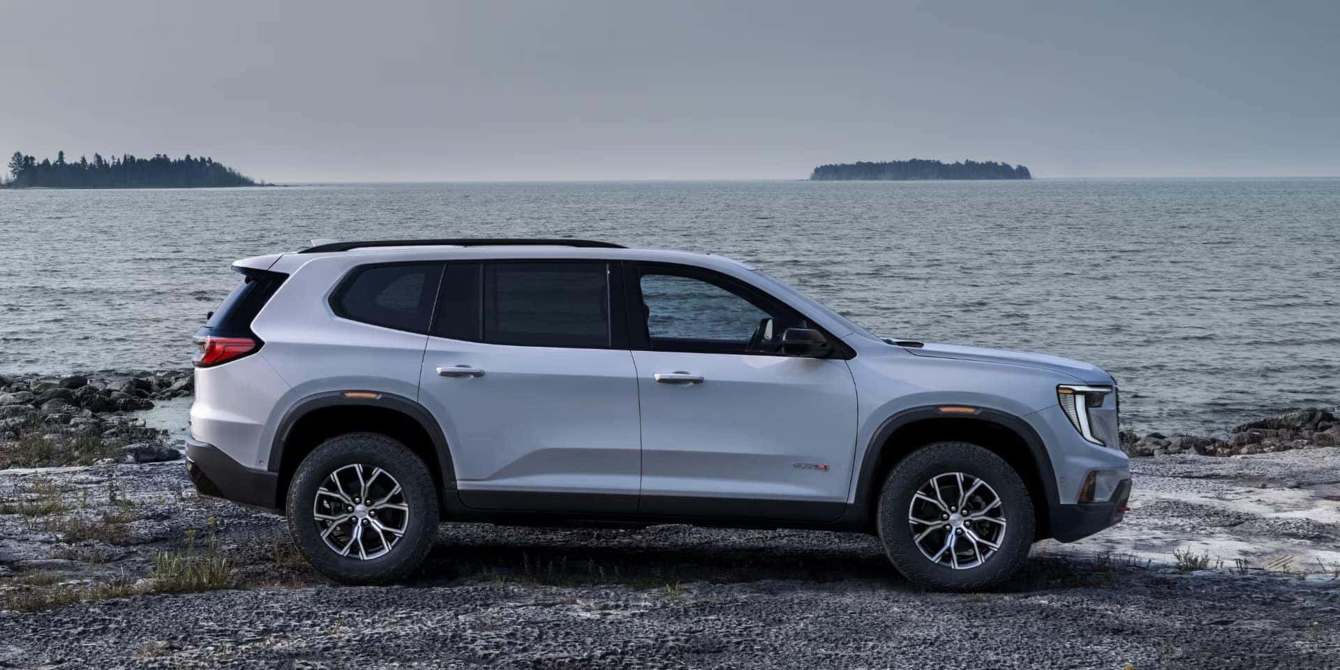 A white SUV is parked on a rocky shoreline with a large body of water and small islands in the background under an overcast sky. The vehicle is viewed from the side, showcasing its profile and sporty design.