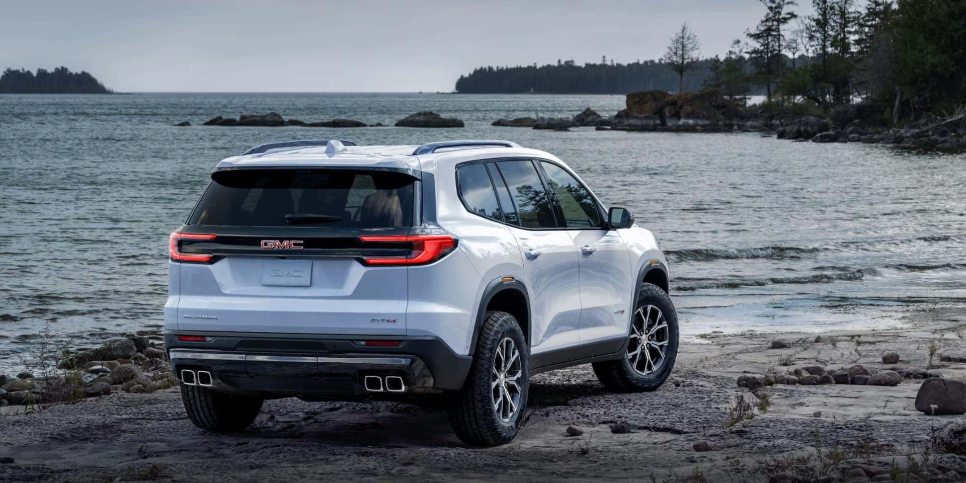 A white SUV is parked on a rocky shoreline next to a body of water. The sky is overcast, and trees are visible on a distant island. The rear of the vehicle is prominently displayed, featuring dual exhaust pipes and a branded emblem.