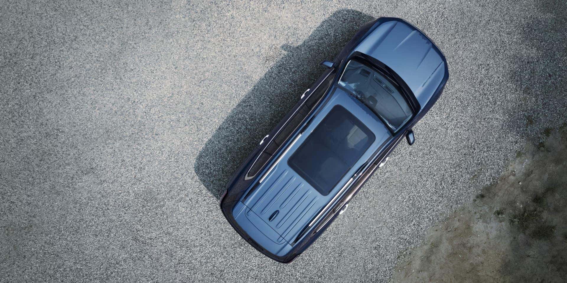Aerial view of a silver car parked on a gravel surface. The car has a panoramic sunroof and sleek lines. The gravel extends to the right, with a small patch of grass visible in the bottom right corner.