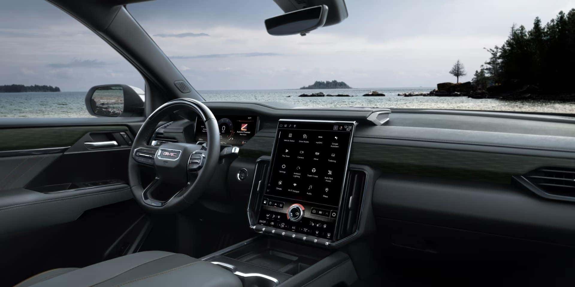 Car interior with a modern dashboard featuring a large touchscreen display and steering wheel with the GMC logo. The vehicle is parked facing a scenic view of a lake with distant islands and a cloudy sky.