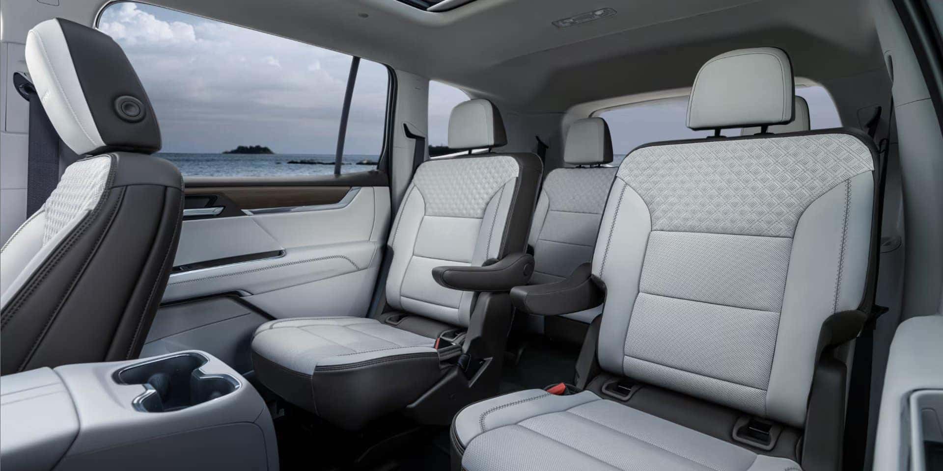 Interior of a modern car showing three spacious, light gray seats with armrests; panoramic windows offer a view of the ocean and cloudy sky, creating a serene ambiance.