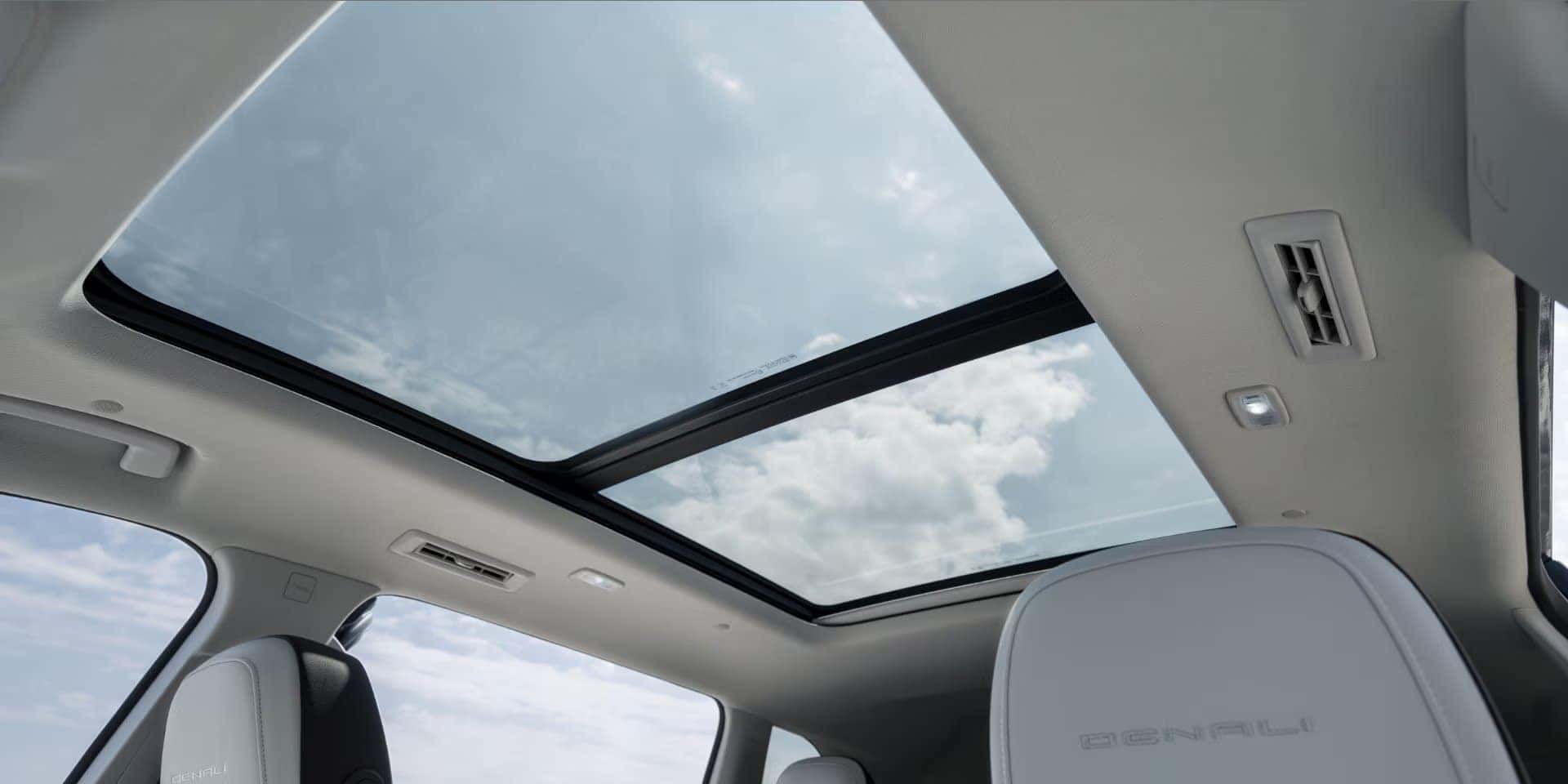 The image shows the interior of a vehicle with a panoramic sunroof. The sky is visible through the glass, with a few clouds. The seats are light-colored and labeled with Denali.