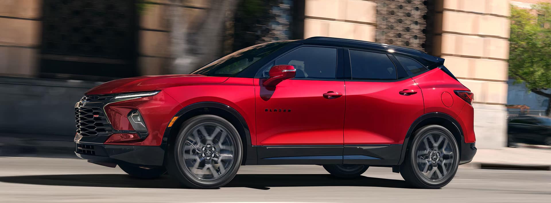 A red SUV with black and chrome accents is driving on a city street. Its a four-door vehicle with large alloy wheels, a sleek design, and tinted windows. The background features blurred buildings and trees.