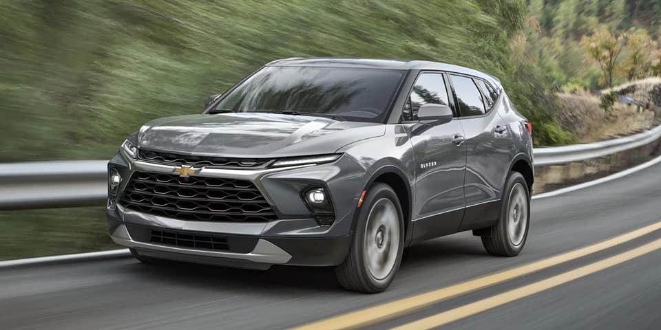 A gray Chevrolet SUV is driving on a two-lane road with yellow lines. The background shows blurred greenery, indicating motion. The SUV has a sleek design with distinctive front grille and headlights.