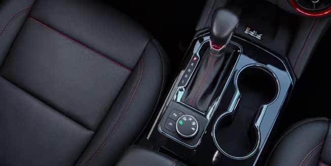 Close-up of a cars center console featuring a gear shift, two cup holders, and controls for driving modes. The interior has black leather with red stitching, giving a sleek and modern appearance.