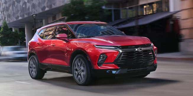 A red SUV is driving on an urban street. The vehicle features sleek, modern design elements with sharp headlights and a prominent grille. Buildings and trees are blurred in the background, suggesting motion.