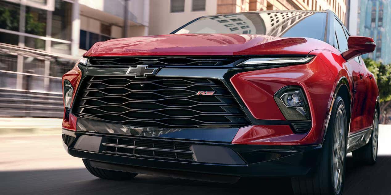 A red Chevrolet car with the RS badge on the front grille is parked on a city street. The image highlights the vehicles sleek design and prominent front emblem, with buildings and trees visible in the background.