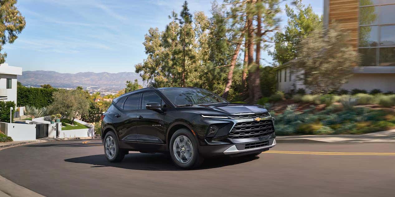 A sleek black SUV driving on a winding road through a residential area with lush greenery and modern homes. Hills and mountains are visible in the background under a clear blue sky.