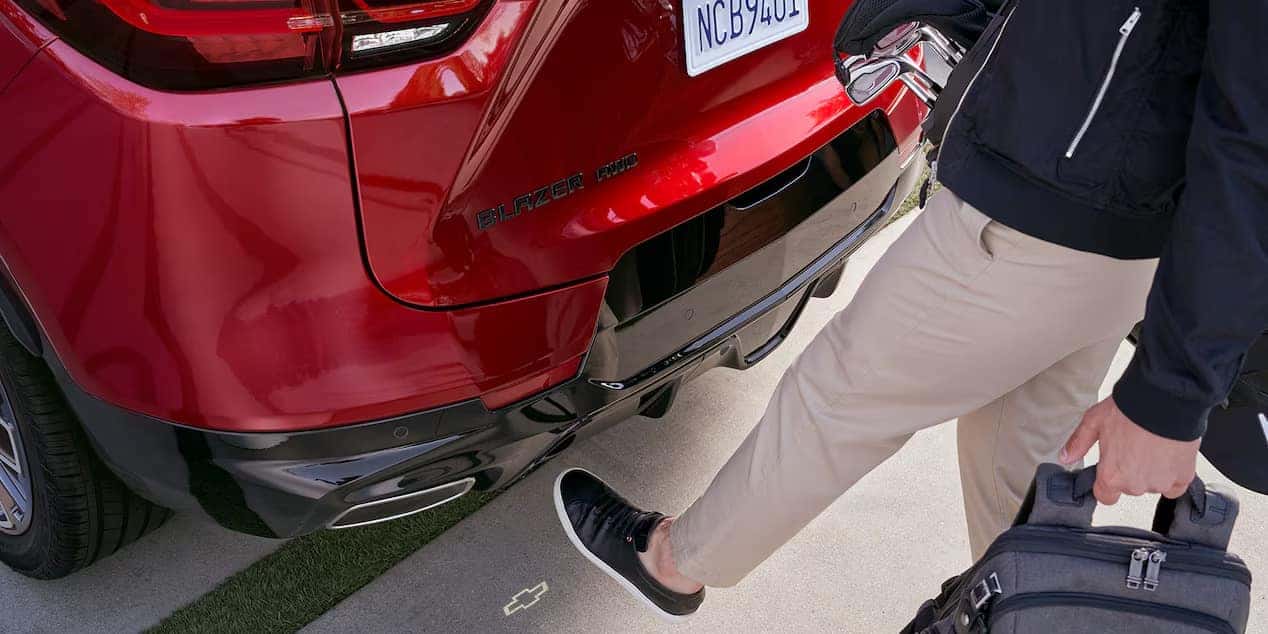 Person using their foot to open the rear hatch of a red Chevrolet Blazer RS. They are wearing beige pants, black sneakers, and holding a black bag. The cars license plate is partially visible.