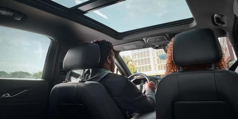 Two people are inside a car with a sunroof, viewed from the back seat. The driver holds the steering wheel, and sunlight fills the space. Buildings and trees are visible through the windows.