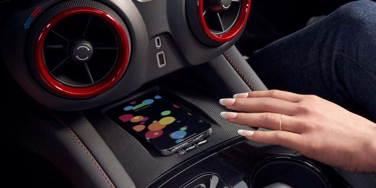 A hand with neatly manicured nails and a gold band rests on a cars center console. A smartphone with a colorful, abstract dots wallpaper is lying nearby. The dashboard features two circular air vents with red trim.