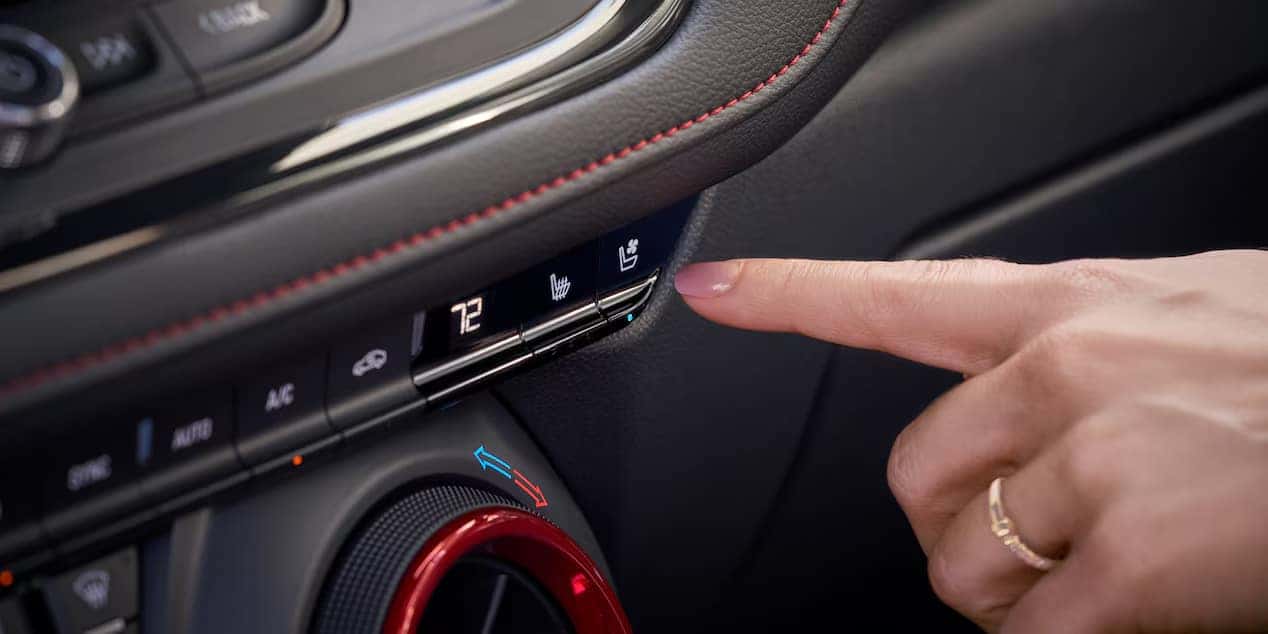 A hand is pressing a button on a cars dashboard to turn on the seat heating function. The dashboard displays include the temperature set to 72 degrees and controls for air conditioning and other features.