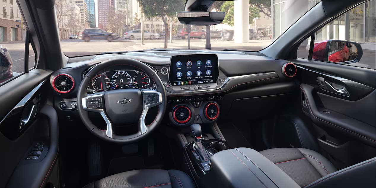 Interior view of a modern car dashboard featuring a multi-function steering wheel, digital display, and touchscreen with various apps. The trim includes red accents on air vents. Urban street scene visible through the windscreen.