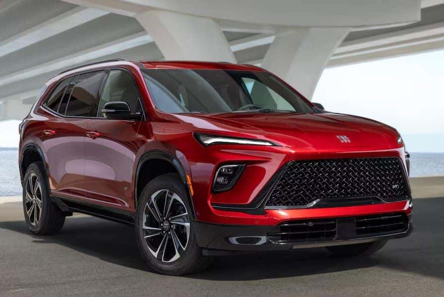A shiny red SUV is parked under a bridge. The vehicle features sleek design lines, a large black grille, and modern headlights. Its windows are tinted, and it rests on a smooth, sunlit surface with a body of water in the background.