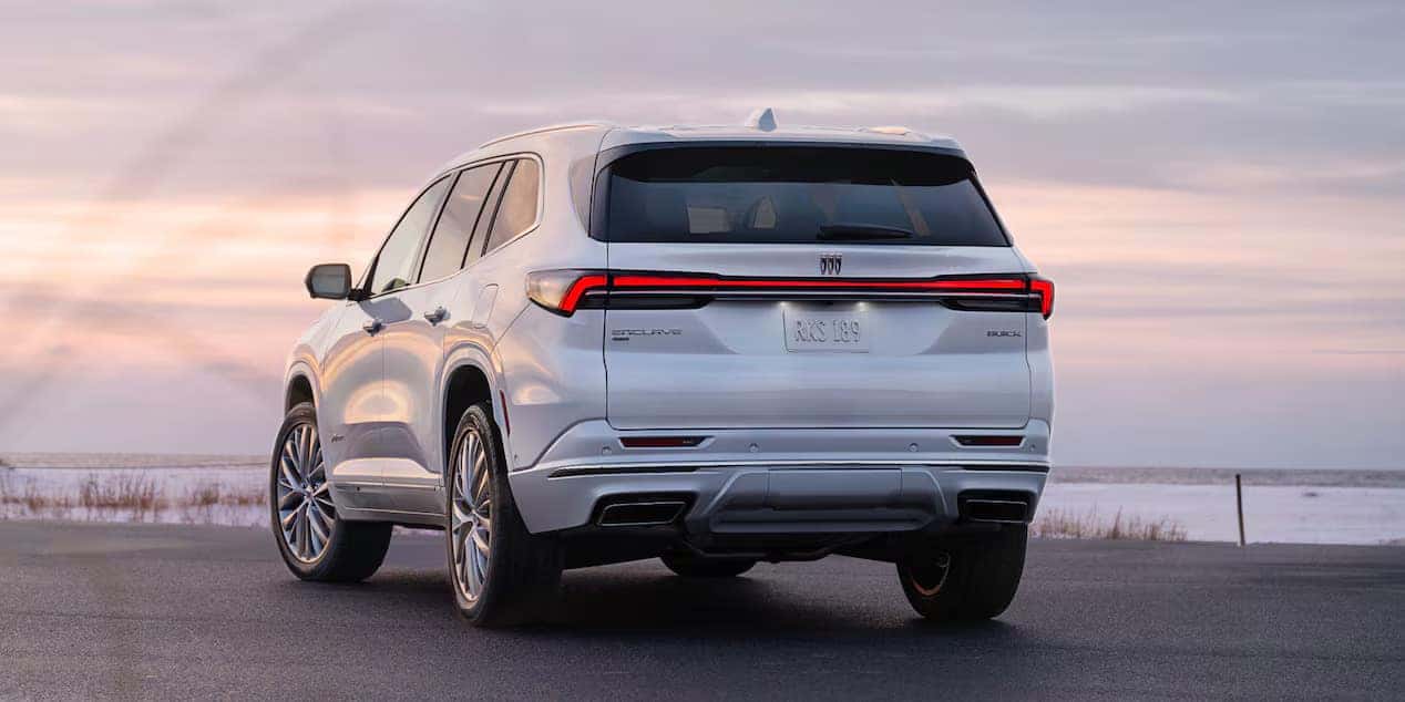 A white SUV is parked on a paved surface with a scenic backdrop of a cloudy sky and a distant shoreline. The vehicles rear design features sleek taillights and a smooth exterior. The sun casts a warm light over the landscape.