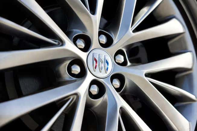 Close-up of a car wheel with a metallic alloy design. The center of the wheel features a round emblem with a tricolor shield. The spokes are sleek and polished, radiating symmetrically from the center.