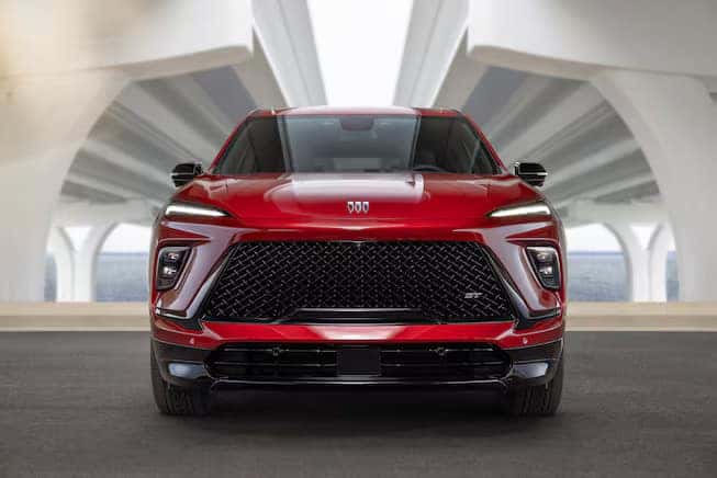 Front view of a sleek, red SUV parked under a modern, architectural structure. The vehicle features a large, black grille and narrow headlights, with a glossy finish reflecting its surroundings.