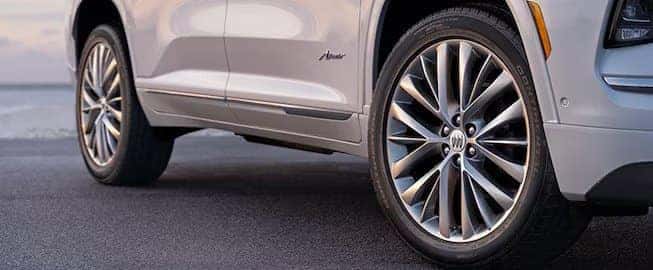 A close-up of the side of a silver SUV on a road, highlighting its sleek, metallic wheels with multi-spoke rims. The cars nameplate is visible on the side fender. The background is blurred, focusing on the vehicles details.