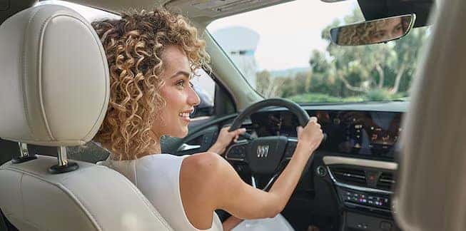 A person with curly hair is smiling while driving a car. The interior features beige leather seats and a modern dashboard. The view outside the car shows greenery, suggesting a sunny day.