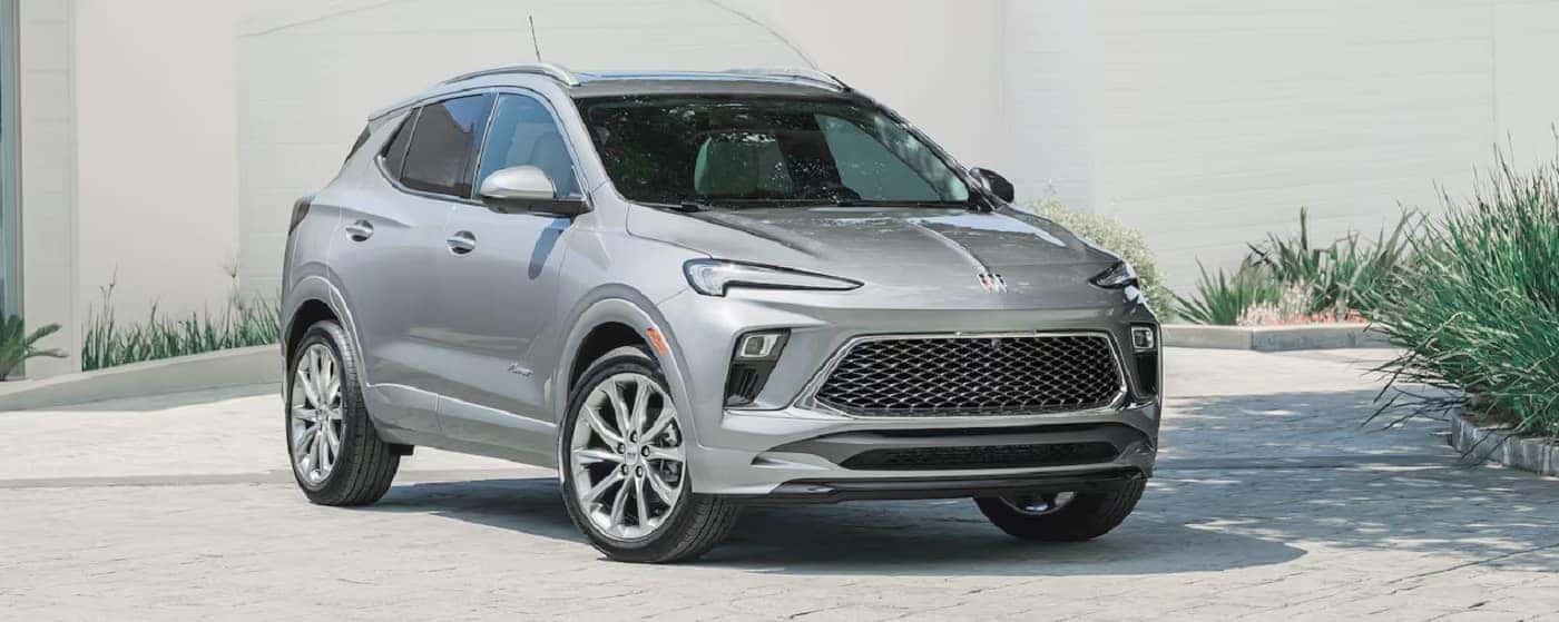 A silver SUV is parked on a paved surface in front of a light-colored wall. The vehicle features a large front grille, sleek headlights, and five-spoke alloy wheels. Green plants are visible in the background.