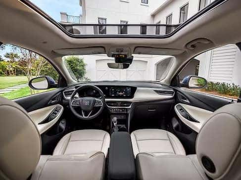 Interior view of a modern car with beige leather seats, a panoramic sunroof, and a sleek dashboard with a touchscreen display. The vehicle is parked in front of a white building with a green lawn visible through the windows.