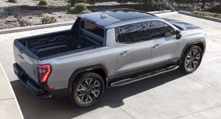 A gray pickup truck parked on a concrete surface, viewed from above and slightly to the side. The truck features a crew cab and a spacious bed. Its design includes sleek, aerodynamic lines and large alloy wheels. Background includes some greenery.