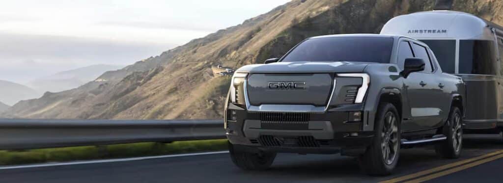 A GMC truck drives on a mountain road, towing an Airstream trailer. The background features rugged hills and a cloudy sky, suggesting a scenic outdoor setting.