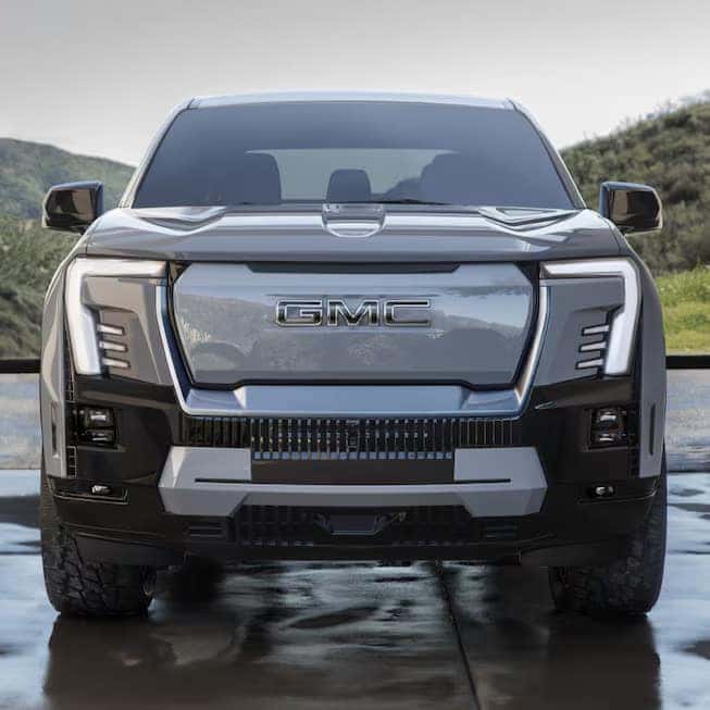 Front view of a shiny, black GMC truck parked on a reflective surface. The vehicle has a prominent grille and sleek LED headlights. Hills and greenery are visible in the background.