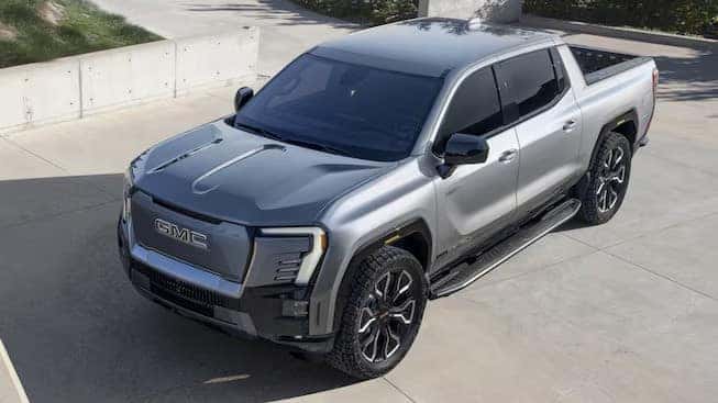 A silver GMC truck parked on a concrete surface. The vehicle features a sleek design with modern headlights and black accents on the grille and wheels. A building with concrete walls is in the background.