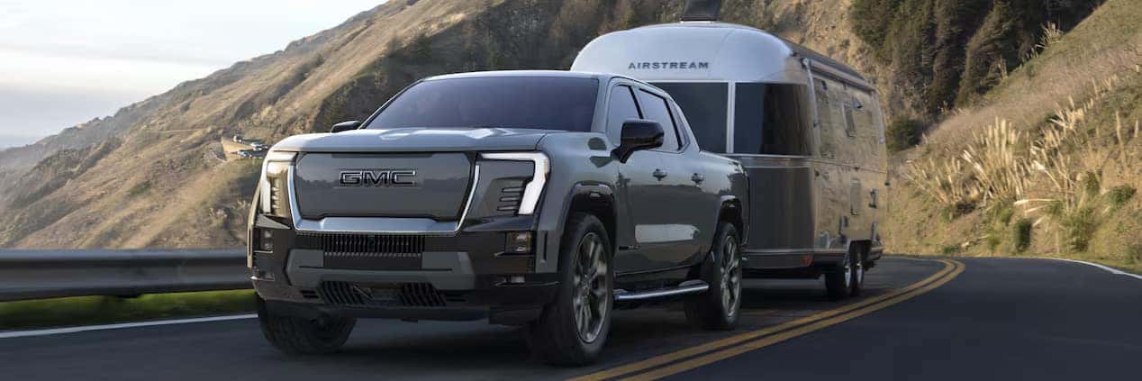 A silver GMC pickup truck is towing a silver Airstream trailer on a scenic coastal road. The background features rocky cliffs and a view of the ocean under a partially cloudy sky.