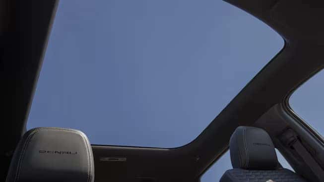 Interior view of a car showcasing a panoramic sunroof. The sky is visible through the large glass roof. Two car seats with stitched upholstery, one with Denali branding, are partially visible.