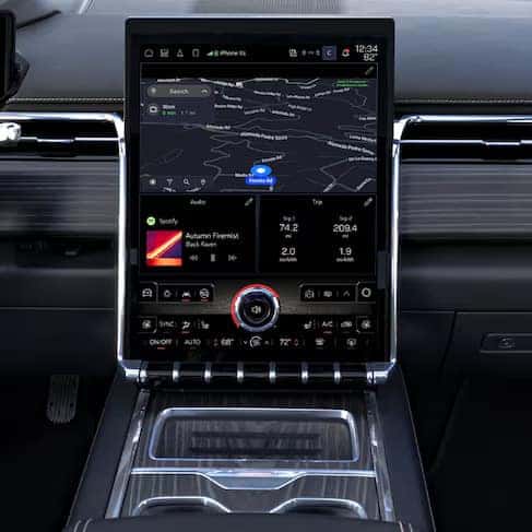 Car infotainment system displaying navigation map, music player, and vehicle information. The interface shows a dark-themed map, audio controls, and climate statistics on a large touchscreen in the center console of a car interior.