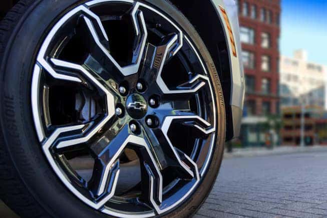 Close-up of a stylish car wheel with a distinctive black and chrome alloy rim featuring a geometric pattern. The Chevrolet logo is visible at the center. The background shows a city street and blurred buildings.