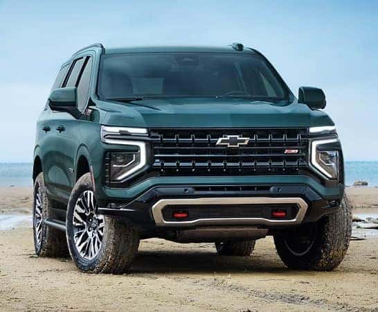 A dark green SUV with a distinctive grille and modern headlights is parked on a sandy beach near the water. The vehicle has bold, off-road tires and a sleek, rugged design, suited for outdoor adventures.