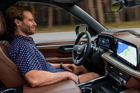 Person with a beard and casual shirt driving a car, looking relaxed. The dashboard displays a digital screen with a navigation map. The interior features brown leather seats. The background shows a blurred outdoor scene.