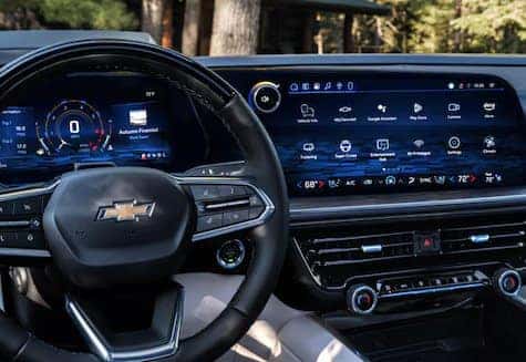 A car dashboard featuring a Chevrolet logo on the steering wheel, with a digital speedometer on the left and a wide touchscreen display on the right, showing various apps and settings, set against a background of trees.