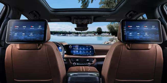 Interior of a car showing two screens on the back of the front seats, displaying various apps. Brown leather seats and a large central dashboard screen are visible. A sunroof reveals a clear sky and outdoor scenery outside the rear window.