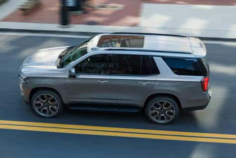 Silver SUV driving on a paved road with yellow double lines. The vehicle is viewed from above, showcasing its roof and side profile. Urban setting with blurred background indicating motion.