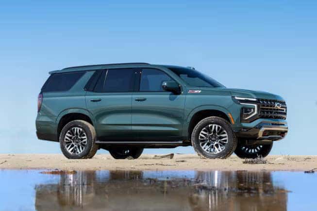 A green SUV is parked on a sandy surface near water. Its a large vehicle with prominent wheel arches and shiny alloy wheels, under a clear blue sky.