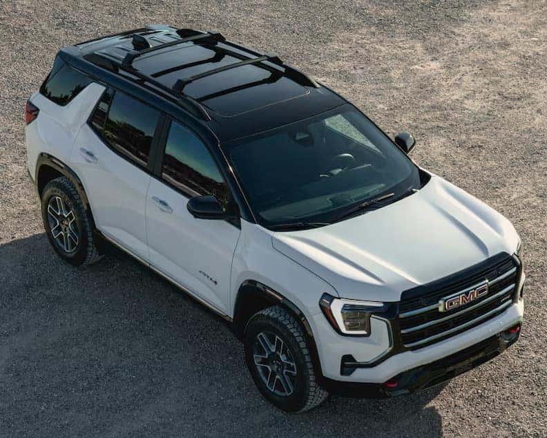 A white GMC SUV is parked on a gravel surface, viewed from a high angle. It features a black roof with sunroofs, prominent front grille, and distinctive wheel rims. The vehicles design highlights a blend of ruggedness and modern style.