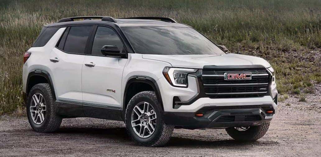 A white GMC AT4 SUV is parked on a gravel path with grass in the background. The vehicle has a bold grille, rugged tires, and black trim accents, giving it a robust appearance. The sky is overcast, adding a muted tone to the setting.